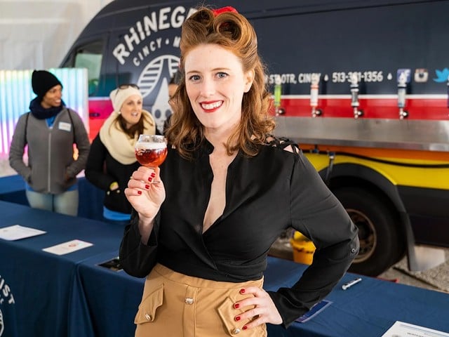 A fair-skinned woman with red hair in victory rolls, wearing red lipstick and smiling while holding a snifter of ale