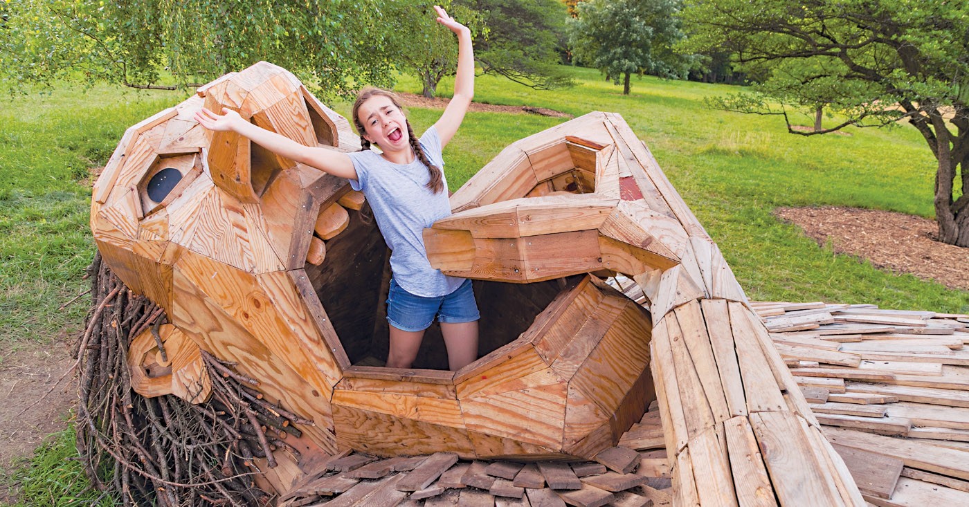 A girl climbs in the mouth of 'Little Artur,' a troll made of reclaimed wood in 'Troll Hunt' by Thomas Dambo, in Bobolink Meadow on the West Side of The Morton Arboretum in Summer. &copy;2018 The Morton Arboretum. All rights reserved. - &copy; Michael Hudson